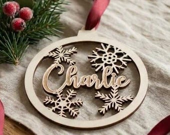 May include: A circular wooden Christmas ornament featuring the name Charlie in script lettering, surrounded by four intricate snowflake cutouts. The ornament is attached to a deep red satin ribbon and accented with a sprig of pine needles.