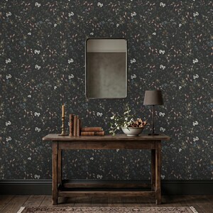 K&ouml;nnte beinhalten: Dunkle florale Tapete mit einem sich wiederholenden Muster aus kleinen wei&szlig;en, blauen und braunen Blumen und Bl&auml;ttern. Ein Holztisch tr&auml;gt B&uuml;cher, eine Lampe, eine Kerze und eine Schale mit Blumen. Ein Spiegel h&auml;ngt an der Wand &uuml;ber dem Tisch.