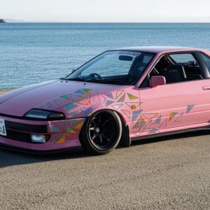 Può includere: Un'auto rosa con un design geometrico sul lato. L'auto è parcheggiata su una spiaggia con il mare sullo sfondo.