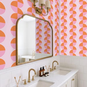 May include: A bathroom with a pink and orange geometric patterned wallpaper. A gold-framed mirror hangs above a white vanity with gold fixtures. The vanity has two sinks, and the room is well-lit.