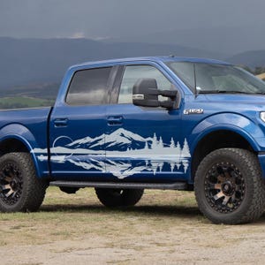 May include: A blue pickup truck with a white mountain and forest decal on the side. The decal features a mountain range, trees, and a sun or moon. The truck has black wheels and is parked on dirt.