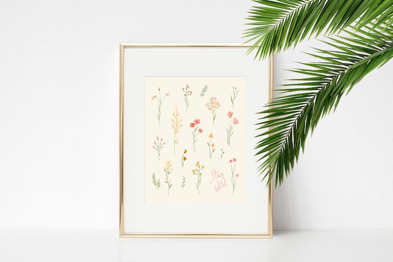 Pu&ograve; includere: Una stampa incorniciata con sfondo bianco e cornice dorata. La stampa presenta una collezione di fiori selvatici disegnati a mano in tonalit&agrave; di rosa, giallo e verde. Il testo "Stay Wild" &egrave; scritto in corsivo rosa nella parte inferiore della stampa.