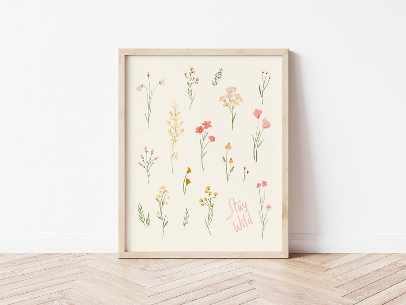 Pu&ograve; includere: Una stampa incorniciata con uno sfondo crema che presenta una collezione di illustrazioni ad acquerello di vari fiori e piante. Il testo "Stay Wild" &egrave; scritto in corsivo rosa nella parte inferiore della stampa.