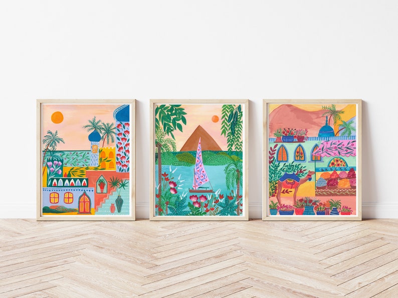 May include: Three framed illustrations of colorful scenes with a Middle Eastern theme. The first illustration shows a building with a blue dome and a pink and orange sunset. The second illustration shows a sailboat on a blue lake with a brown mountain in the background. The third illustration shows a camel walking past a building with a blue dome and a pink and orange sunset.