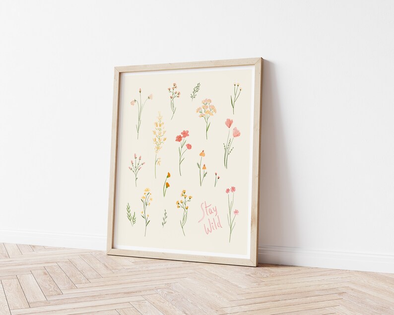 Pu&ograve; includere: Una stampa incorniciata con uno sfondo beige che presenta una collezione di fiori selvatici disegnati a mano in tonalit&agrave; di rosa, giallo e verde. Il testo "Stay Wild" &egrave; scritto in rosa nella parte inferiore della stampa.