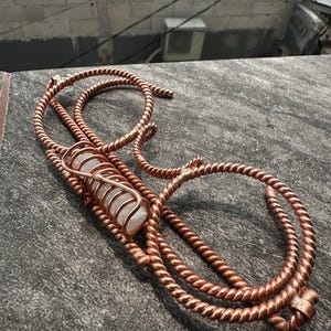 May include: A decorative object crafted from twisted copper wire, featuring a central cylindrical clear quartz crystal encased within a spiral copper wire cage, with the wire extending into curved, rope-like loops and ends.