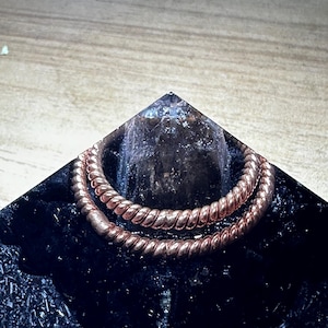 Puede incluir: Una pulsera de metal retorcido de color cobre rodea una estructura cristalina oscura sobre una base negra en forma de pirámide. La textura de la pulsera es visible y la base tiene una superficie texturizada. La estética general es moderna y geométrica.
