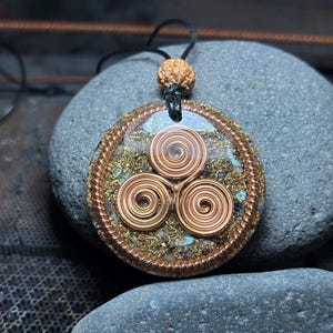 Puede incluir: Un colgante circular con tres espirales de alambre de cobre dispuestas en forma triangular, incrustadas en un disco de resina transparente relleno de virutas metálicas doradas y pequeños fragmentos de piedra azul, bordeado por alambre de cobre trenzado.