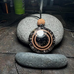 May include: A round pendant necklace with a black center, copper wire spirals, and a small, round, brown bead. The pendant is on a black cord and rests on a gray stone. The pendant has a clear, shiny, circular element.