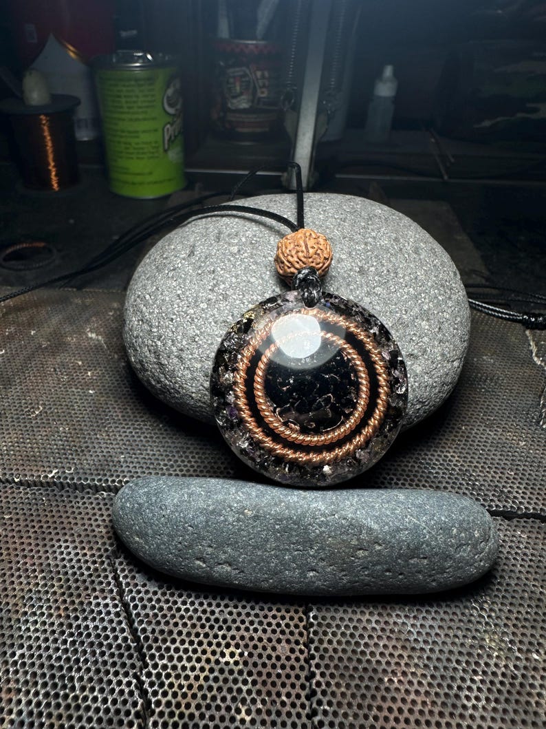 May include: A round pendant necklace with a black center, copper wire spirals, and a small, round, brown bead. The pendant is on a black cord and rests on a gray stone. The pendant has a clear, shiny, circular element.