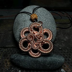May include: Copper-colored pendant necklace with a floral design. The pendant is made of woven copper wire and is suspended from a black cord. A small, textured bead is at the top of the pendant. The necklace rests on a gray stone.