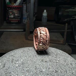 May include: A rose gold ring with a textured, chain-like design. The ring is made of multiple bands that are woven together.