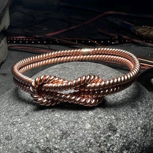 May include: A copper bracelet formed from two strands of twisted wire, featuring a central knot design. The metallic surface has a polished, reflective finish, and the two strands are joined together at the back with a smooth, silver-toned metal clasp.