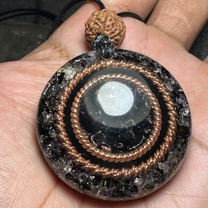 May include: A round pendant with a black center and copper wire spiral design. The pendant is set in a matrix of dark, sparkling material and is suspended from a black cord. A brown, textured bead sits above the pendant.