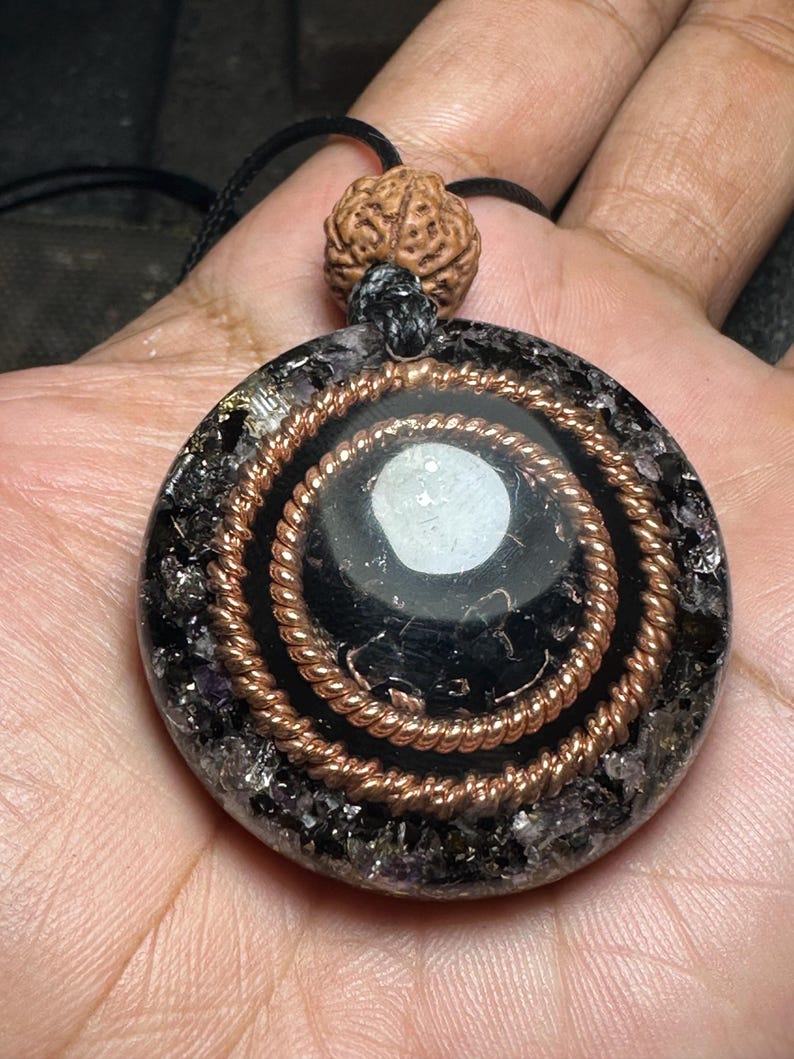 May include: A round pendant with a black center and copper wire spiral design. The pendant is set in a matrix of dark, sparkling material and is suspended from a black cord. A brown, textured bead sits above the pendant.