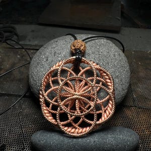 Op de afbeelding: Een koperen draad Flower of Life hanger ketting met een bruine houten kraal op een zwart koord.