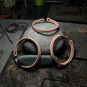 May include: Three copper wire bracelets, each with a braided design, are arranged on a gray stone and a black metal surface. The bracelets are handmade and have a rustic, organic look.