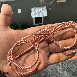 May include: A handcrafted copper wire accessory featuring a twisted rope texture and a central spiral cage element. The metallic structure is formed into two circular loops connected by a coiled section that holds a clear, faceted crystal point.