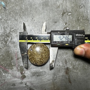 May include: A round, gold-colored pendant with a small hole, measured at 4.246 mm using a digital caliper. The pendant has a textured surface and is set against a metallic background.