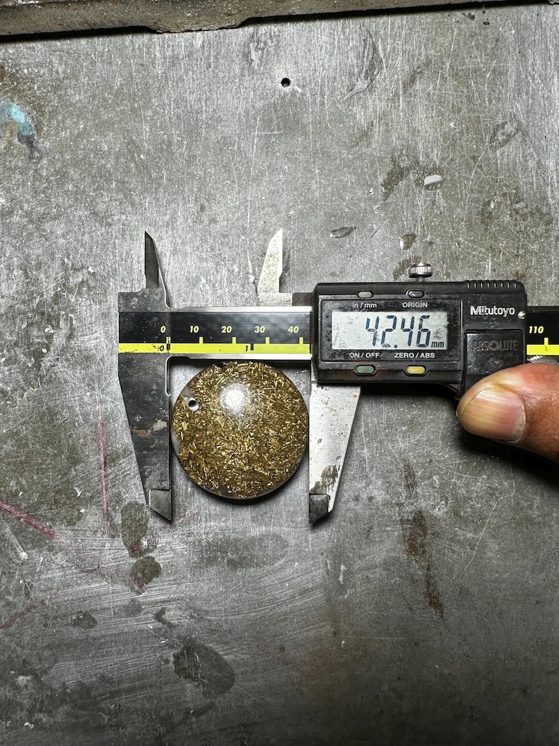 May include: A round, gold-colored pendant with a small hole, measured at 4.246 mm using a digital caliper. The pendant has a textured surface and is set against a metallic background.