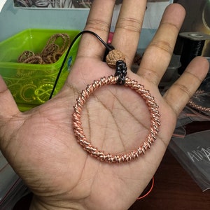 May include: A copper wire ring with a twisted design, attached to a black cord with a brown wooden bead.