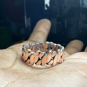 May include: A copper ring with a unique, interlocking design. The ring is made of multiple small, rounded pieces that fit together to create a continuous band.