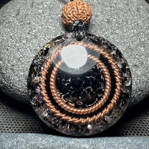 May include: Round pendant with a black center, copper spiral, and a brown bead. The pendant is on a black cord. The pendant is made of resin and contains small crystals. The background is a gray stone.
