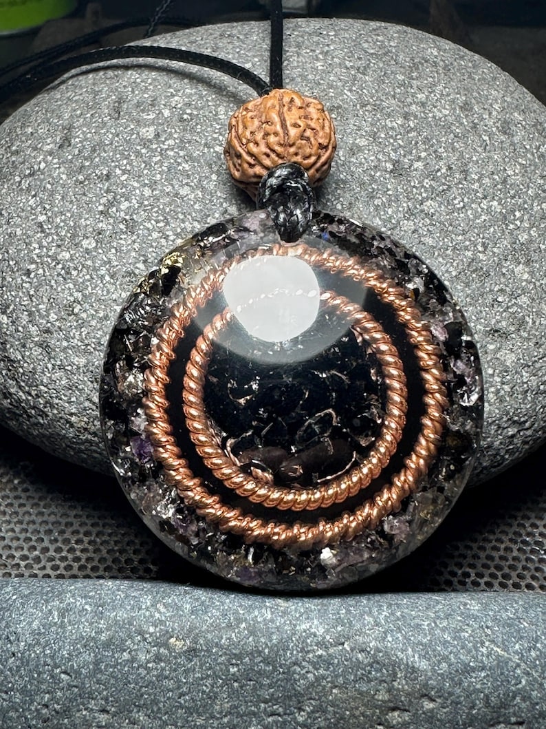 May include: Round pendant with a black center, copper spiral, and a brown bead. The pendant is on a black cord. The pendant is made of resin and contains small crystals. The background is a gray stone.