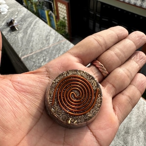 May include: A circular resin pendant featuring a prominent copper wire spiral embedded on the top surface. The base of the pendant contains small metallic shavings and flecks suspended within the clear, hardened resin material.