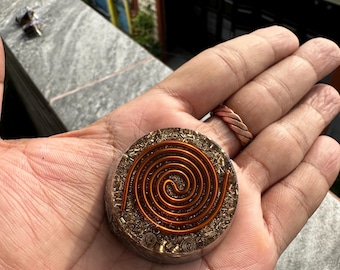 May include: A circular resin pendant featuring a prominent copper wire spiral embedded on the top surface. The base of the pendant contains small metallic shavings and flecks suspended within the clear, hardened resin material.