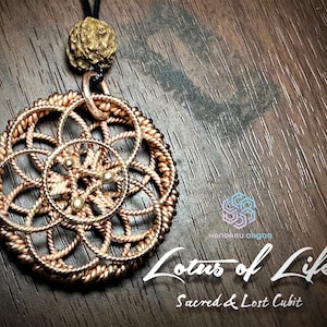 May include: A gold-toned metal pendant with a circular design featuring a geometric pattern. The pendant is hanging from a black cord and has a small, dark brown bead at the top. The pendant is labeled "Lotus of Life Sacred & Lost Cubit".