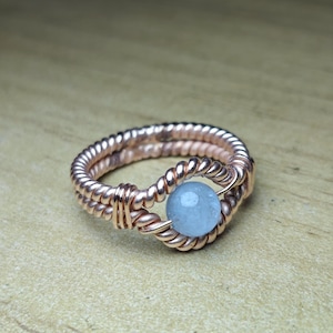 May include: A handcrafted copper wire ring featuring a light blue gemstone. The ring's band is intricately woven, creating a textured appearance. The gemstone is round and centrally placed, adding a pop of color to the copper design.