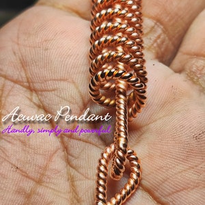 Może przedstawiać: Miedziany wisiorek Acuac z motywem skręconej liny. Wisiorek jest trzymany w dłoni i zawiera tekst "Acuac Pendant Handly, simply and powerful" oraz "Empowerment & Lost Cubit".