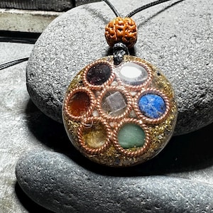 Peut inclure: Un collier pendentif artisanal avec un pendentif circulaire en résine contenant des pierres précieuses colorées. Le pendentif est encadré de fil de cuivre et suspendu à un cordon noir. Une perle marron texturée se trouve en haut du collier. Le pendentif mesure environ 4 cm de diamètre.