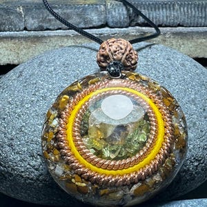Puede incluir: Un colgante redondo de cristal marrón y dorado con un diseño de alambre de cobre. El colgante está en un cordón negro y está rodeado de piedras.