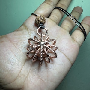 May include: A copper-colored pendant necklace featuring a spherical, openwork design. The pendant is suspended from a black cord and includes a natural-looking bead at the top. The intricate metalwork creates a unique, eye-catching accessory.