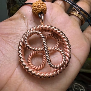 May include: A copper-colored pendant necklace featuring a circular design with intertwined rope-like elements. The pendant is suspended from a black cord and is topped with a brown, textured bead. The jewelry is held in a hand.