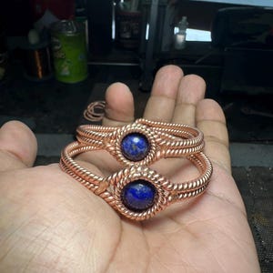 Puede incluir: Dos pulseras envueltas en alambre de cobre con una piedra de lapislázuli azul en el centro de cada una.