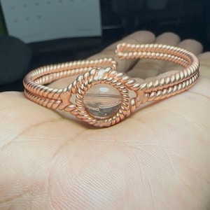 May include: Copper-coloured wire bracelet with a central circular design. The bracelet features a woven rope-like pattern and a clear, round centrepiece with a landscape image. The bracelet is displayed on a hand.