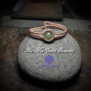 Puede incluir: Un brazalete de alambre de cobre con una piedra preciosa cabujón verde. El brazalete está sobre una piedra gris con el texto "Ra-Mu Cabis Bracelet" y "Handaru Orgon" impreso en ella.