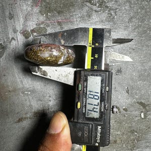 May include: A clear, oval-shaped stone with gold and brown inclusions is measured with a digital caliper. The caliper displays a measurement of 18.74 mm. The stone is on a metal surface.