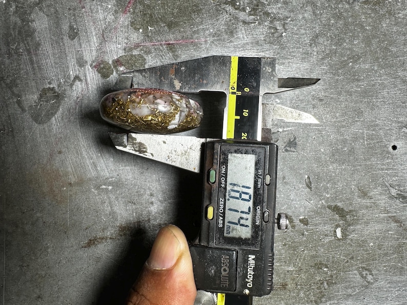 May include: A clear, oval-shaped stone with gold and brown inclusions is measured with a digital caliper. The caliper displays a measurement of 18.74 mm. The stone is on a metal surface.
