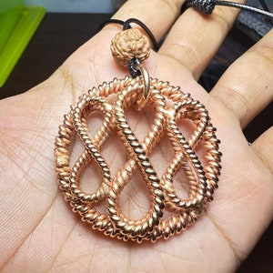 Könnte beinhalten: Eine goldfarbene Metallkette mit einem runden, filigranen Anhänger. Der Anhänger besteht aus gedrehtem Draht und hat eine geknotete Schnur.