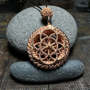 Op de afbeelding: Een koperen draad hanger ketting met een geometrisch ontwerp. De hanger is rond met een bloemvormig patroon in het midden. De ketting hangt aan een bruin leren koord.