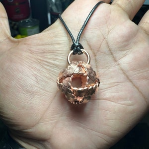 Può includere: Una collana con un ciondolo a sfera intrecciata di colore rame. Il ciondolo è appeso a un cordoncino nero.