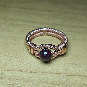 May include: A handcrafted copper ring with a dark red, round gemstone. The ring features a twisted wire design around the band and a wire wrap holding the gemstone in place. The ring is a unique piece of jewelry.