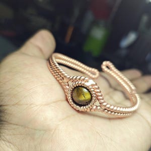 May include: Copper cuff bracelet featuring a central, round tiger's eye stone. The bracelet has a twisted rope design around the stone and along the band. The warm tones of the copper and stone create a natural, earthy aesthetic.