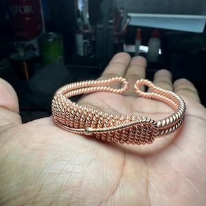 Op de afbeelding: Een koperkleurige armband met een spiraalvormig draadontwerp. De armband heeft een centraal gedeelte van strak gewikkeld draad, geflankeerd door gedraaide draadbanden. Een kleine, ronde kraal accentueert het ontwerp. De armband wordt op een hand getoond.
