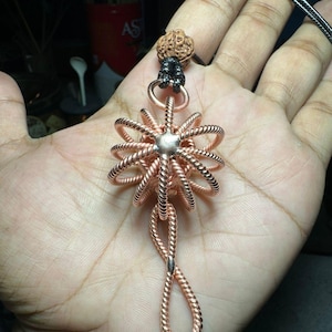 Puede incluir: Un colgante de alambre de cobre con un diseño en forma de estrella y un lazo anudado en la parte inferior. El colgante está unido a un cordón negro con una cuenta de madera marrón.