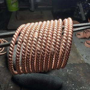 May include: A stack of 10 copper bracelets with a twisted rope design. The bracelets are stacked on top of each other and are in focus. The background is out of focus.